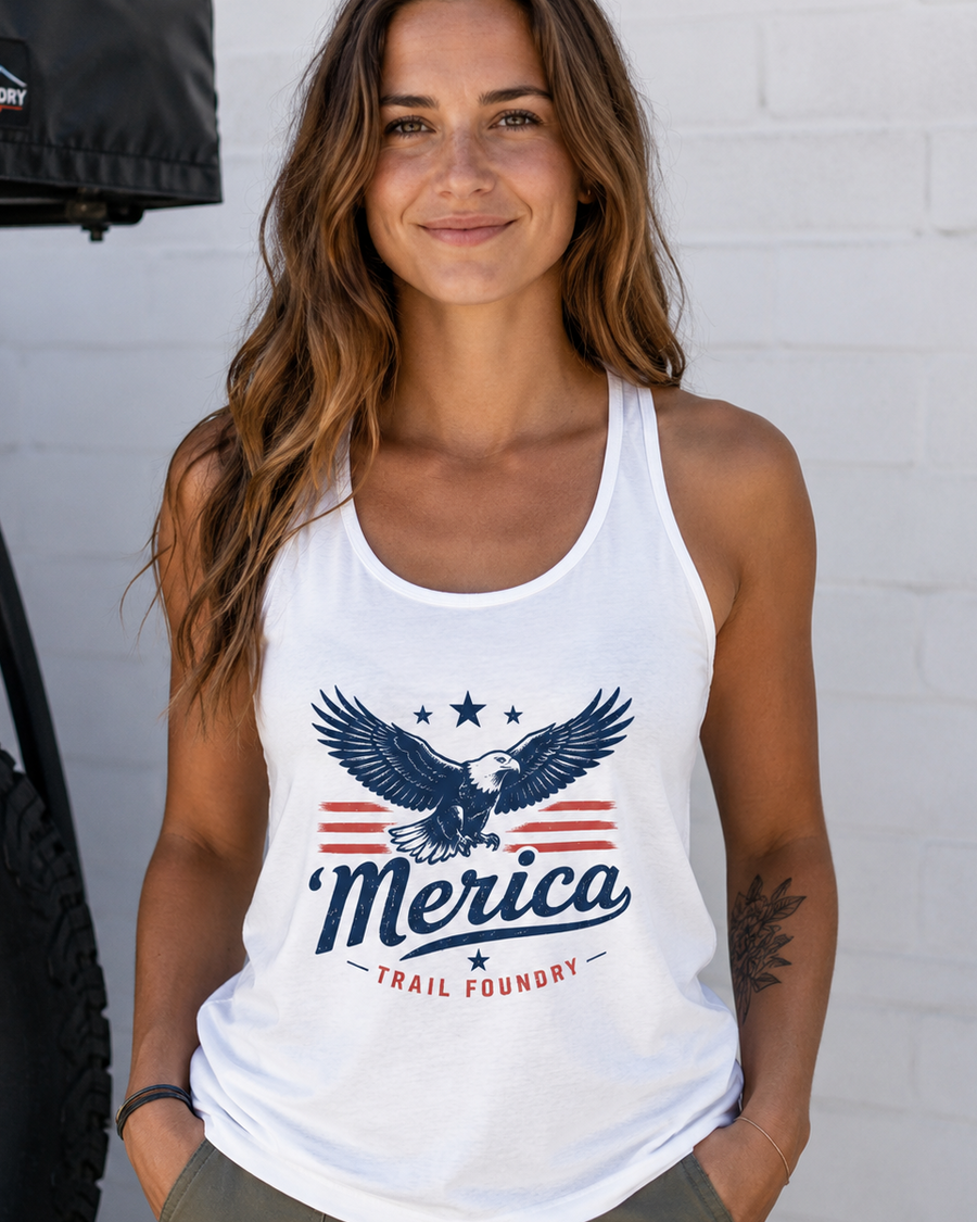Woman wearing a white tank top with 'Merica' and eagle design, standing next to a vehicle.