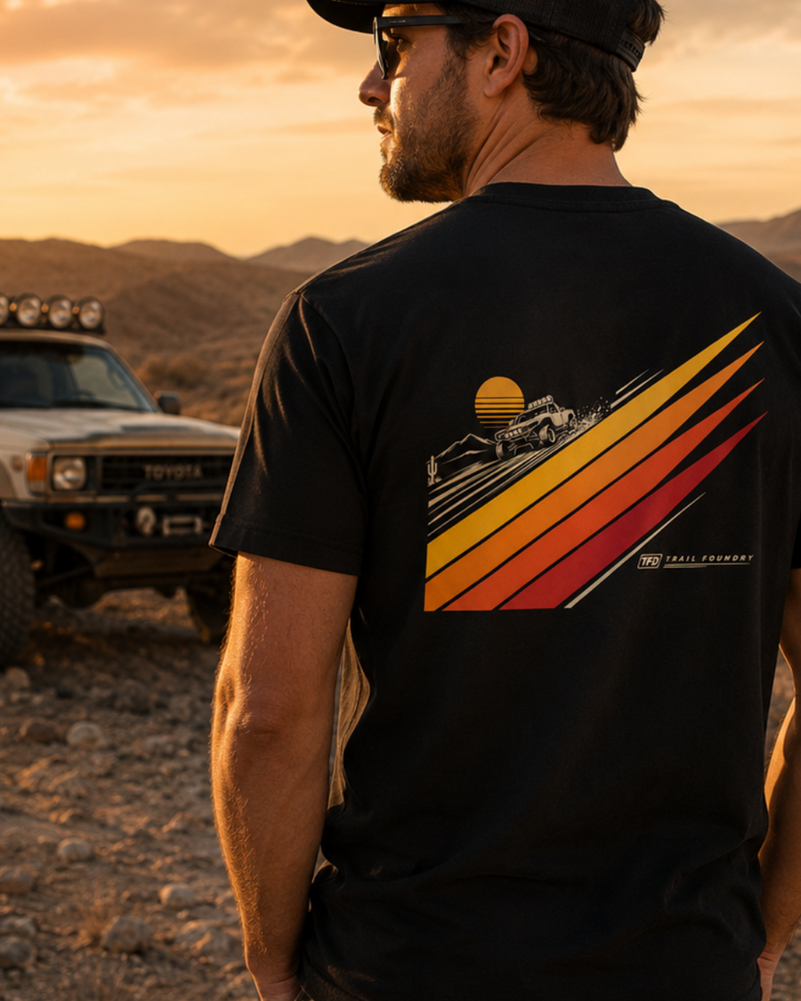 Man wearing a black t-shirt with a graphic design in a desert landscape with a vehicle.